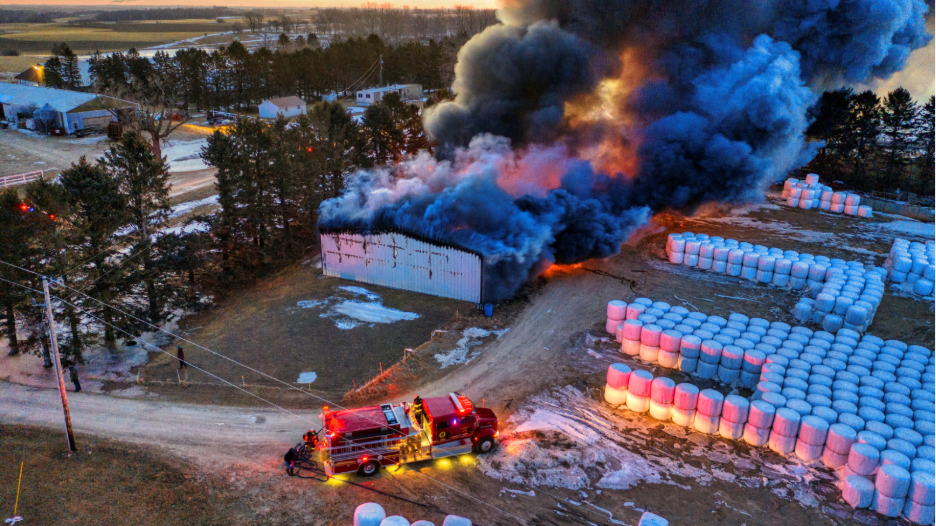 A red fire truck with lights on is parked on a dirt road near a burning white barn emitting thick black smoke at dusk, surrounded by trees and stacks of wrapped hay bales.