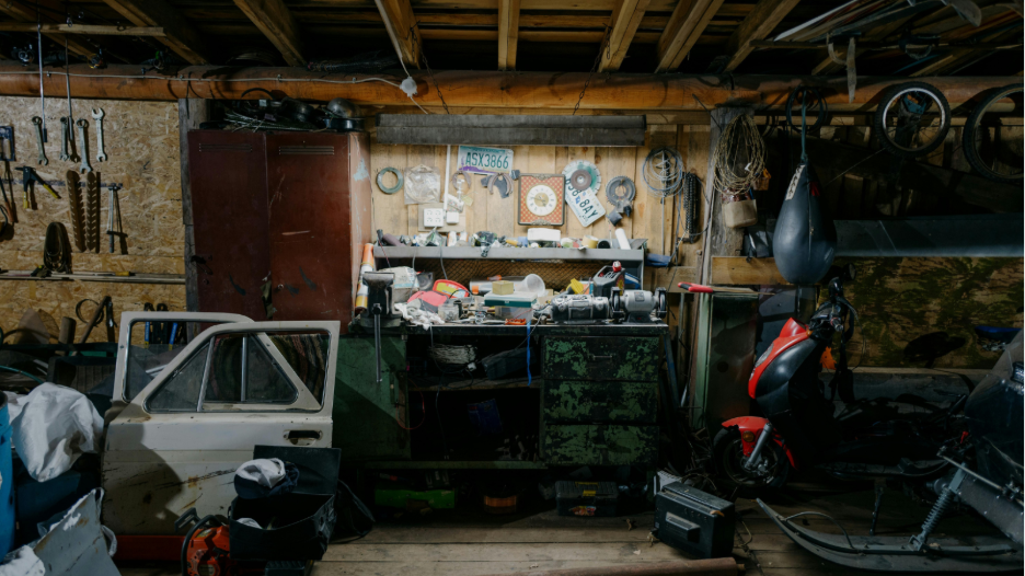 Dimly lit cluttered workshop with wooden beams overhead, a white vehicle door leaning against a green workbench covered with tools and parts, a red scooter parked nearby, various tools hanging on the wall, a red metal cabinet, coiled wires, and a black punching bag.