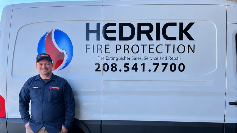 A man wearing a navy blue Hedrick Fire Protection uniform and cap stands smiling in front of a white van with the Hedrick Fire Protection logo and contact number 208.541.7700.