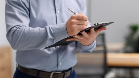 Person in a blue buttoned shirt writing on a clipboard with a pen, standing in an office setting with blurred chairs and desk in the background.