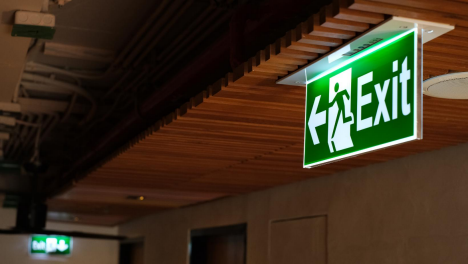 Green illuminated exit sign with a running figure pointing left, mounted on a wooden ceiling indoors.