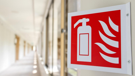 Red and white fire extinguisher sign mounted on a wall in a bright corridor with glass panels.