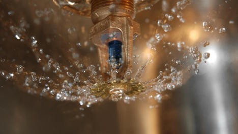 Close-up of a fire sprinkler head spraying water droplets in a circular pattern against a blurred background.