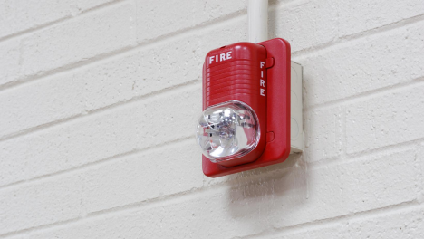 Red fire alarm with a clear strobe light mounted on a white brick wall.