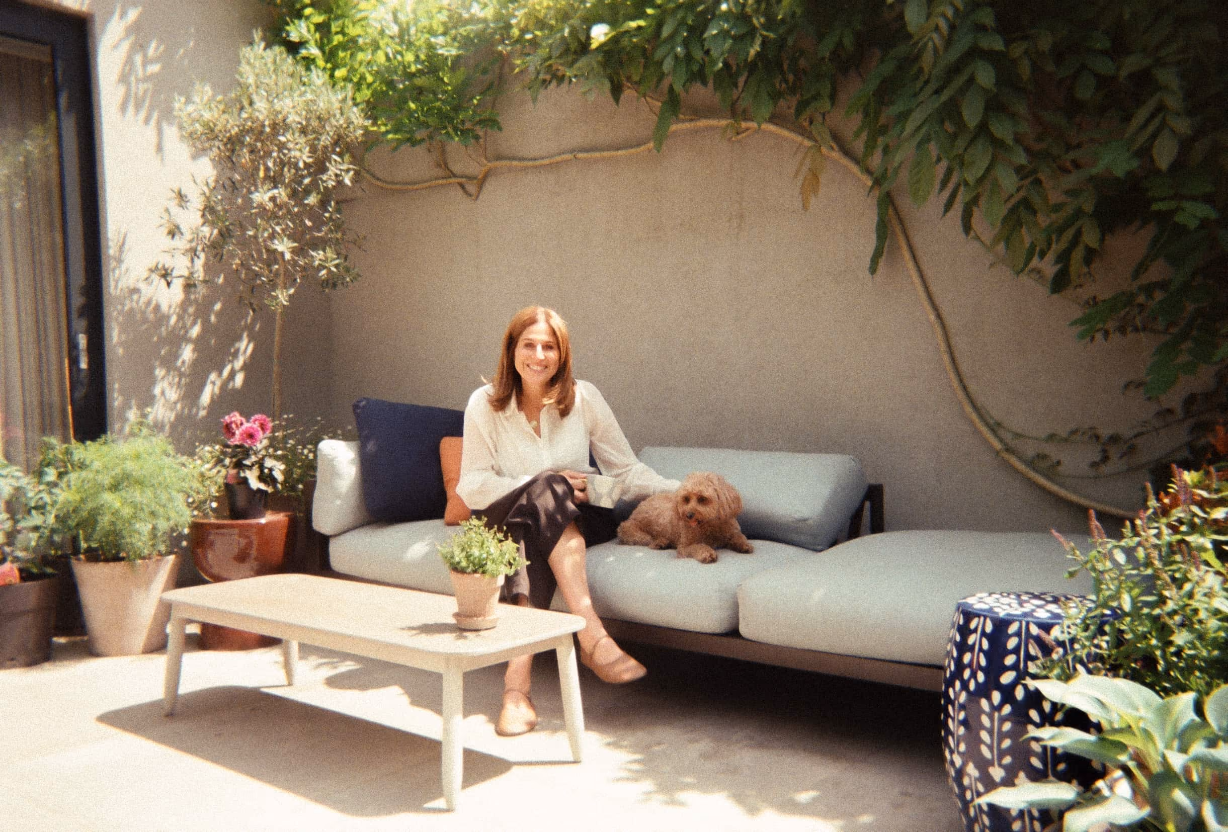 Suzi sitting on outdoor couch petting small dog surrounded by plants and a coffee table.