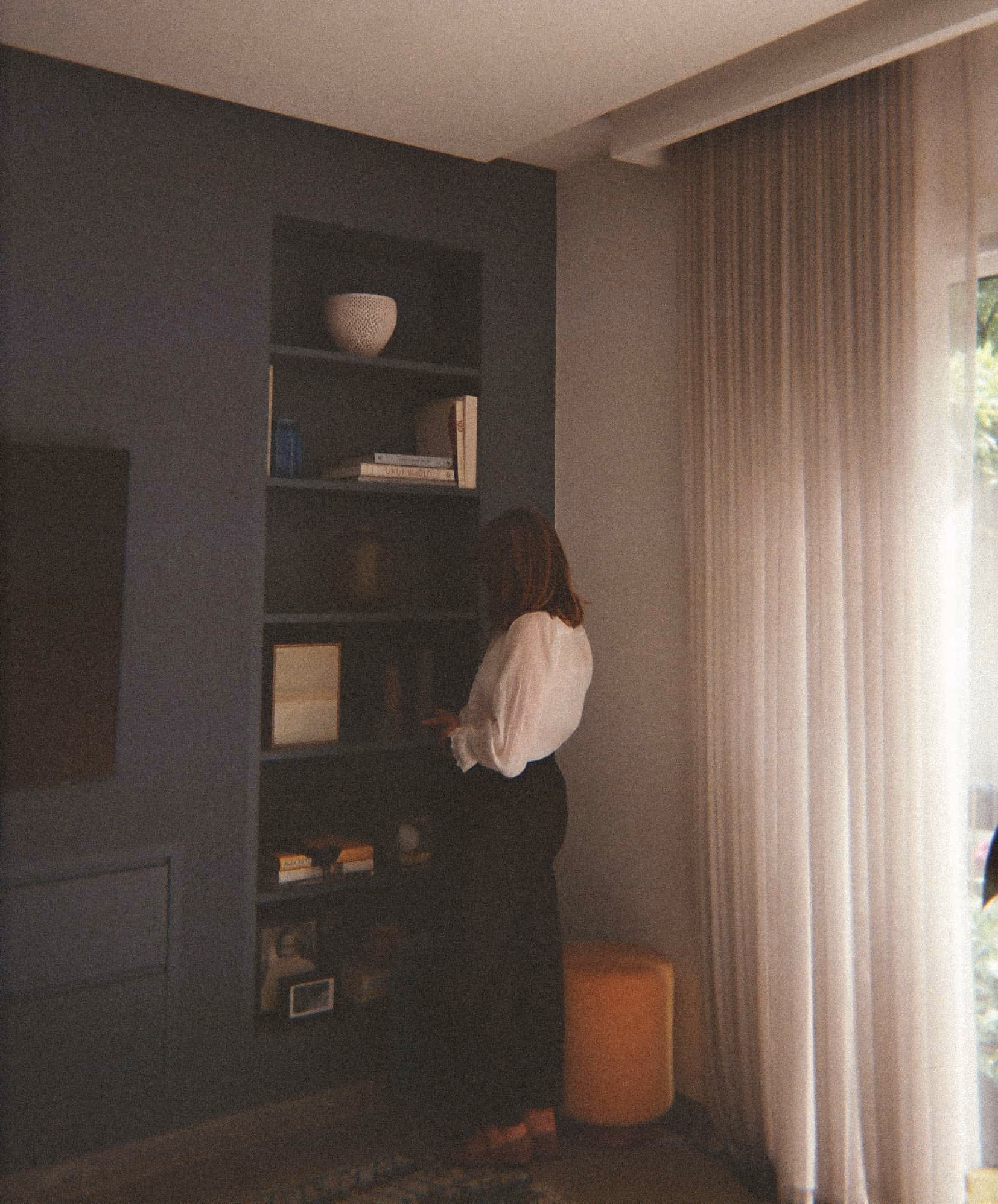 Suzi in white blouse and black skirt browsing items on dark built-in shelves in a softly lit room.