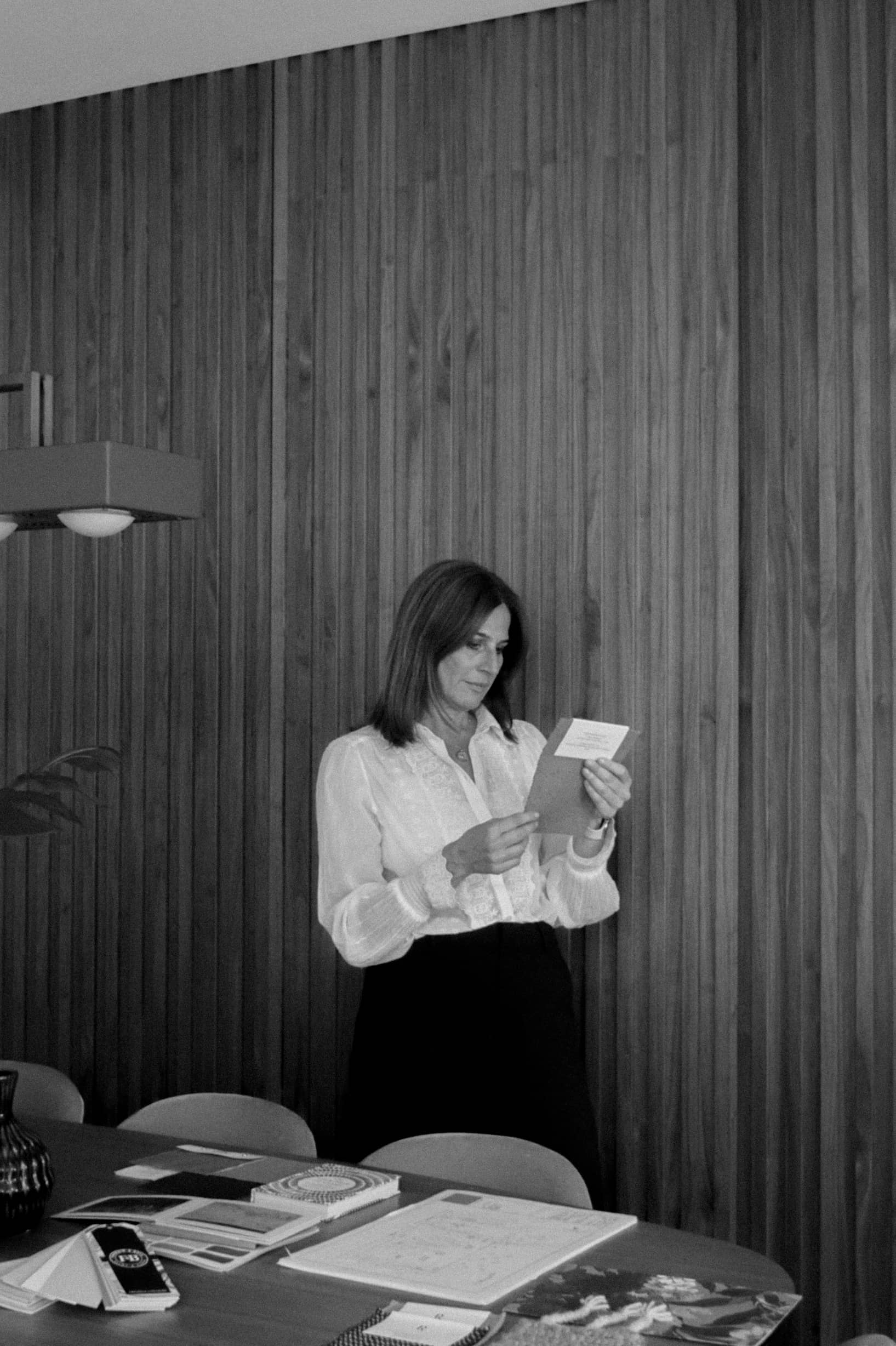 Suzi in white blouse stands reading a card at a table with papers and samples, wooden wall background.