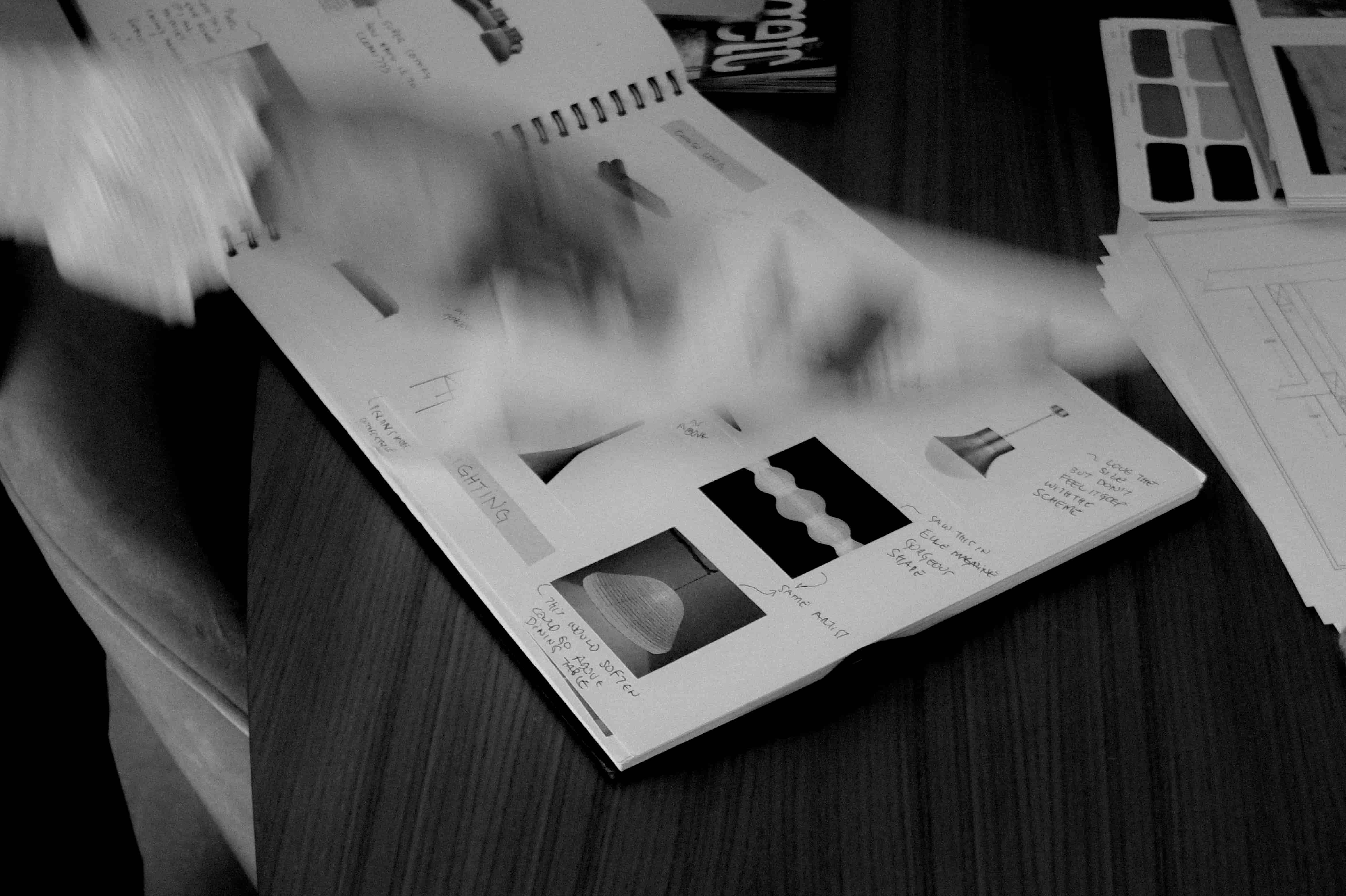 Blurred hand flipping through a notebook with sketches and notes on lighting designs on a wooden table.