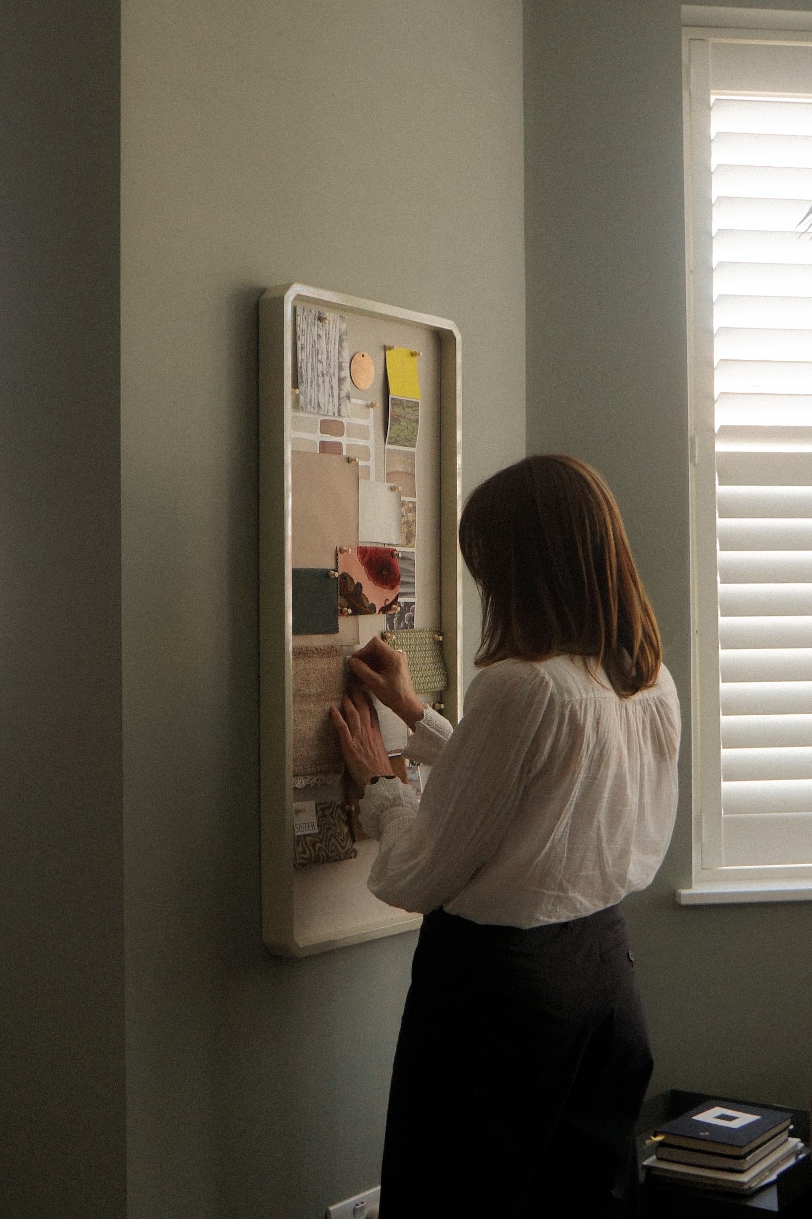 Suzi pinning fabric samples and notes on a wall-mounted board near a window with blinds.
