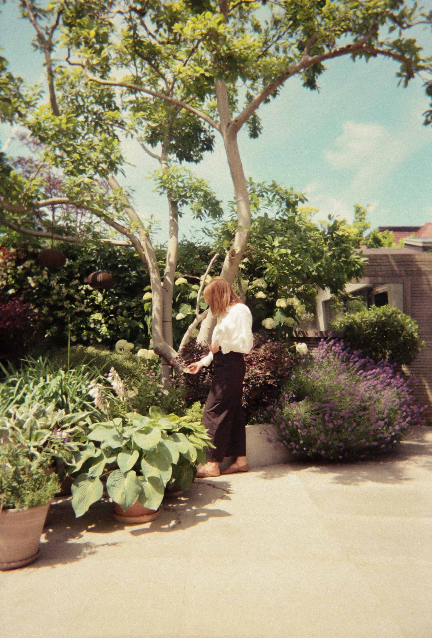Suzi in white sweater and black pants tending plants and flowers in a sunny garden patio.