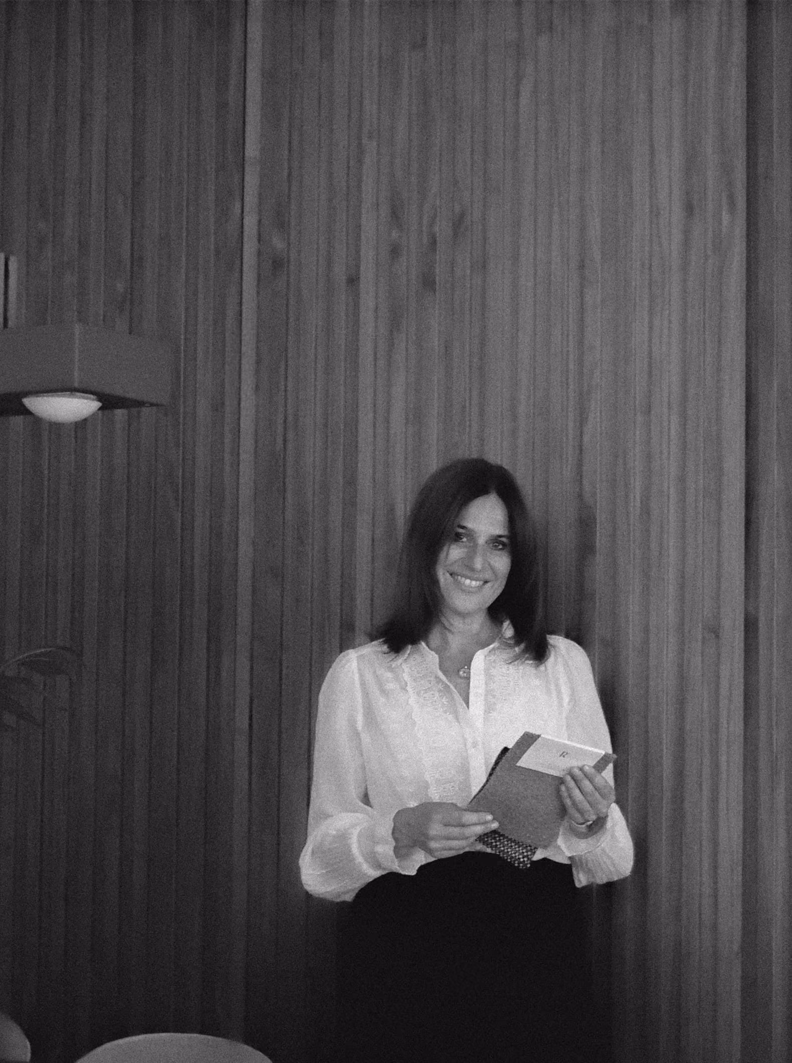 Suzi in a white blouse holding a notebook against a wooden wall.