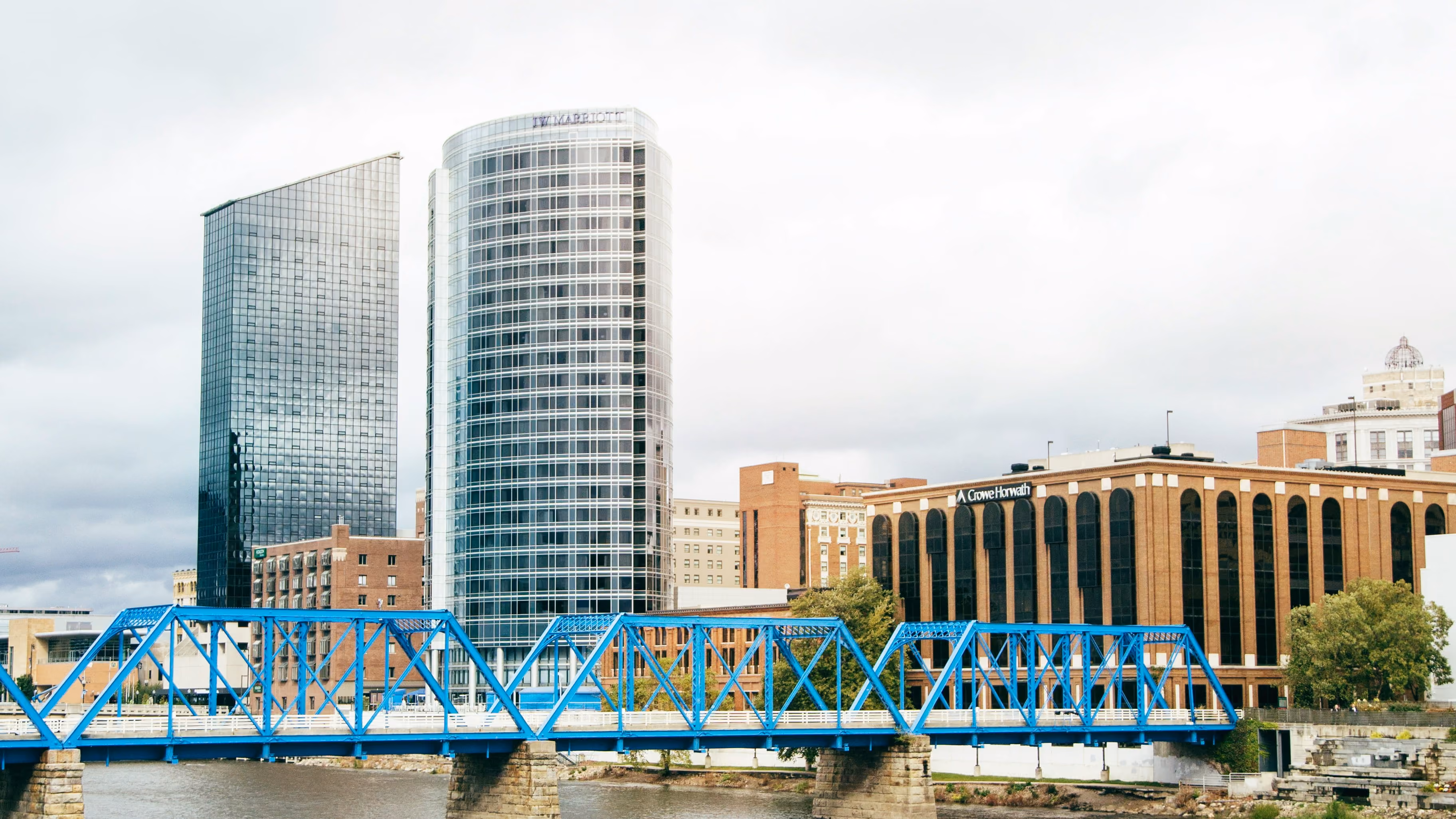 Blue steel truss bridge over a river with modern glass-covered skyscrapers and older brick buildings in the background under a cloudy sky.