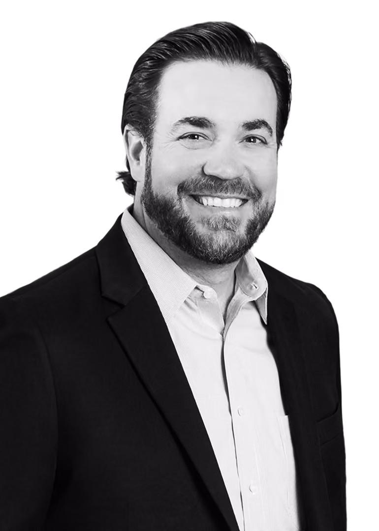 Smiling man with dark hair and beard wearing a light dress shirt under a dark blazer on a black background.
