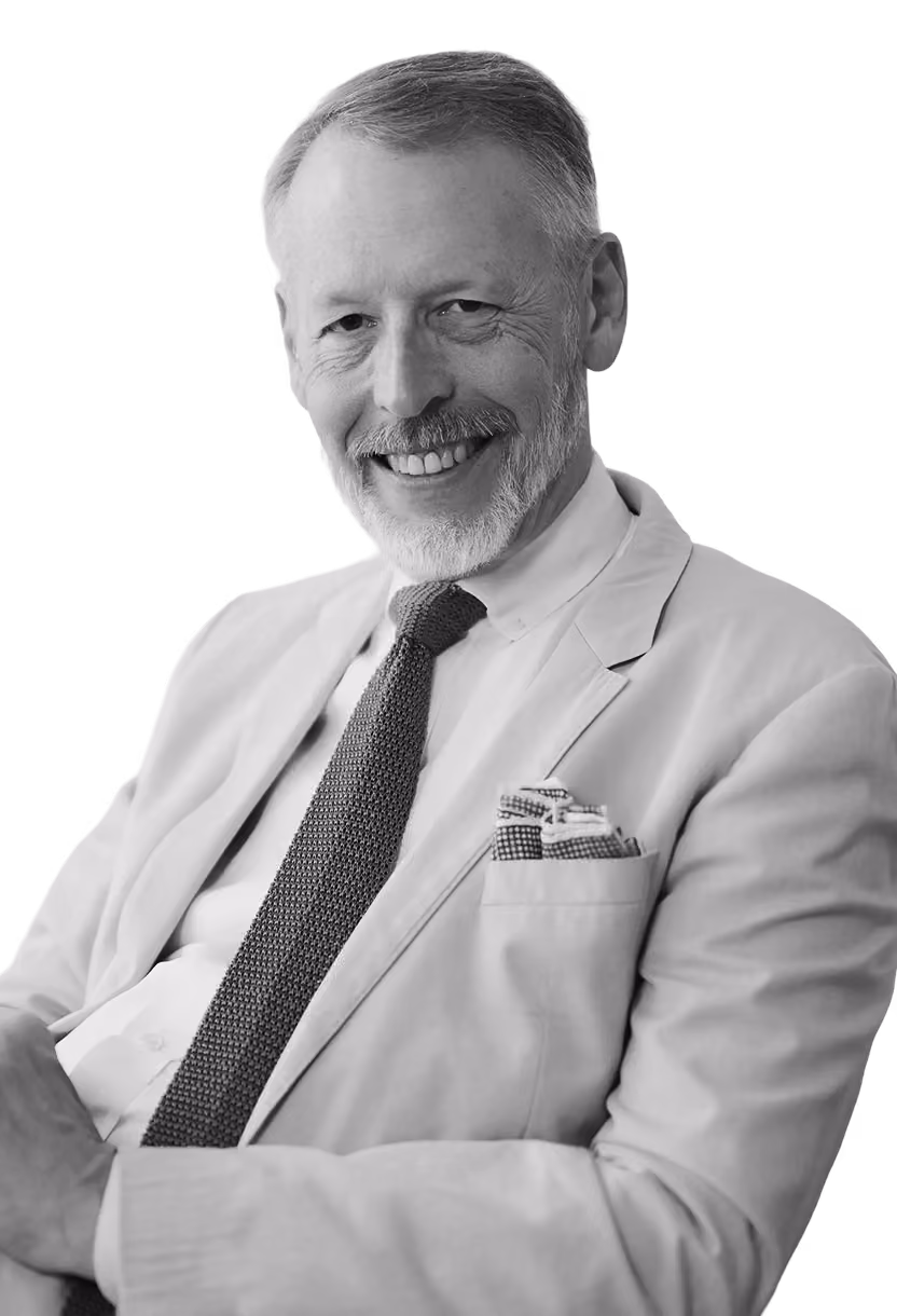 Smiling older man with white hair and beard wearing a light-colored suit jacket, patterned tie, and pocket square, posed with arms crossed.