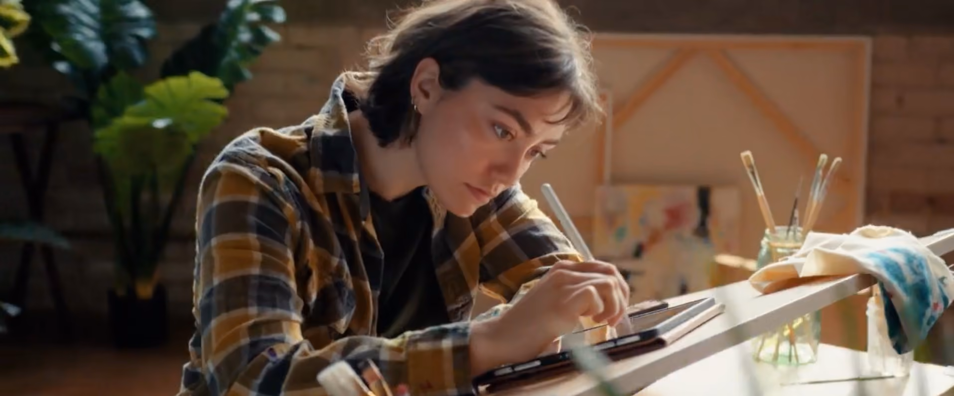 Focused young woman in a plaid shirt using a stylus to draw on a digital tablet on a white table with paintbrushes in jars nearby.