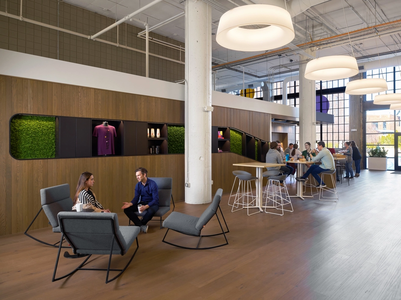 Modern office lounge with wooden floors and walls, featuring green moss panels and display shelves with products. Two people sit and talk in gray rocking chairs in the foreground, while a group of people chat and work at high tables and stools near large windows letting in natural light.
