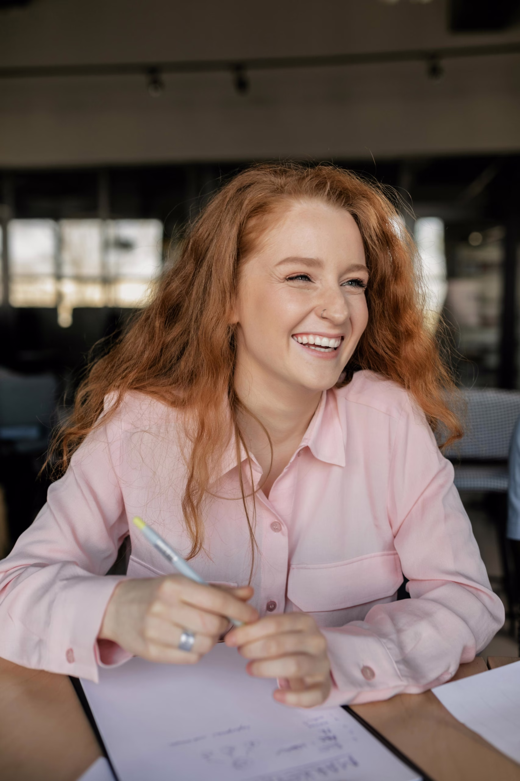A red-haired woman wearing a pink shirt is smiling and holding a pen while sitting at a table with papers in a modern office.