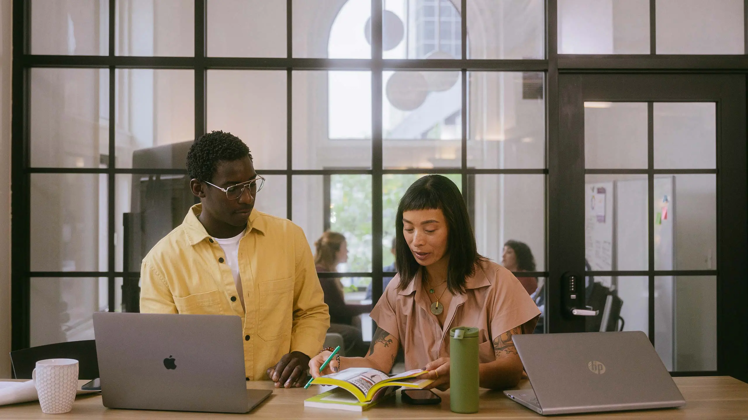 Two people working together at a table with laptops and a book in a modern office with glass walls and blurred colleagues in the background.