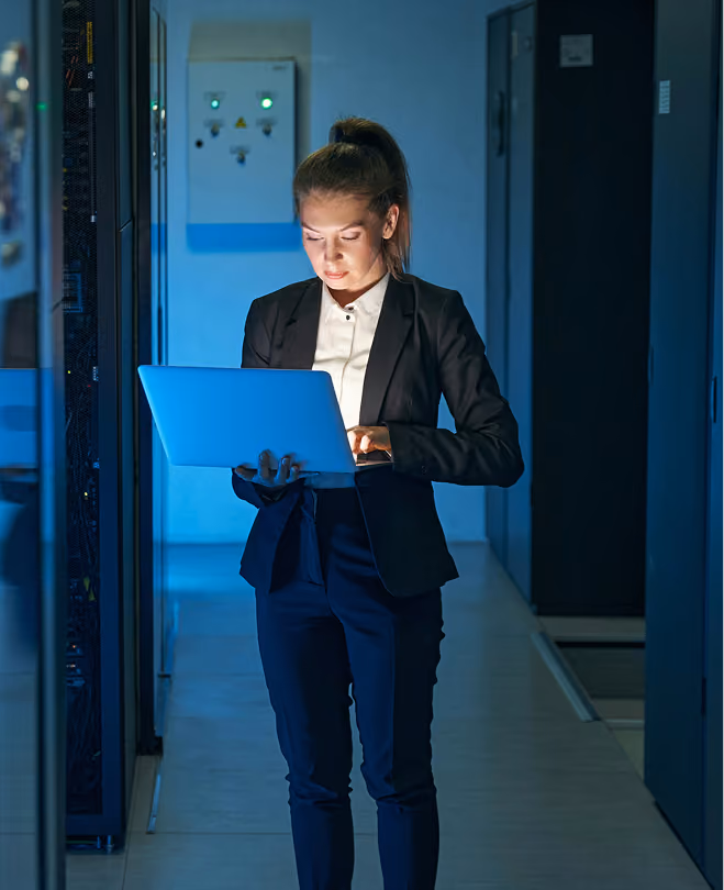 Woman in business attire standing in a dimly lit data center corridor, holding and working on a laptop with blue light illuminating her face and surroundings.