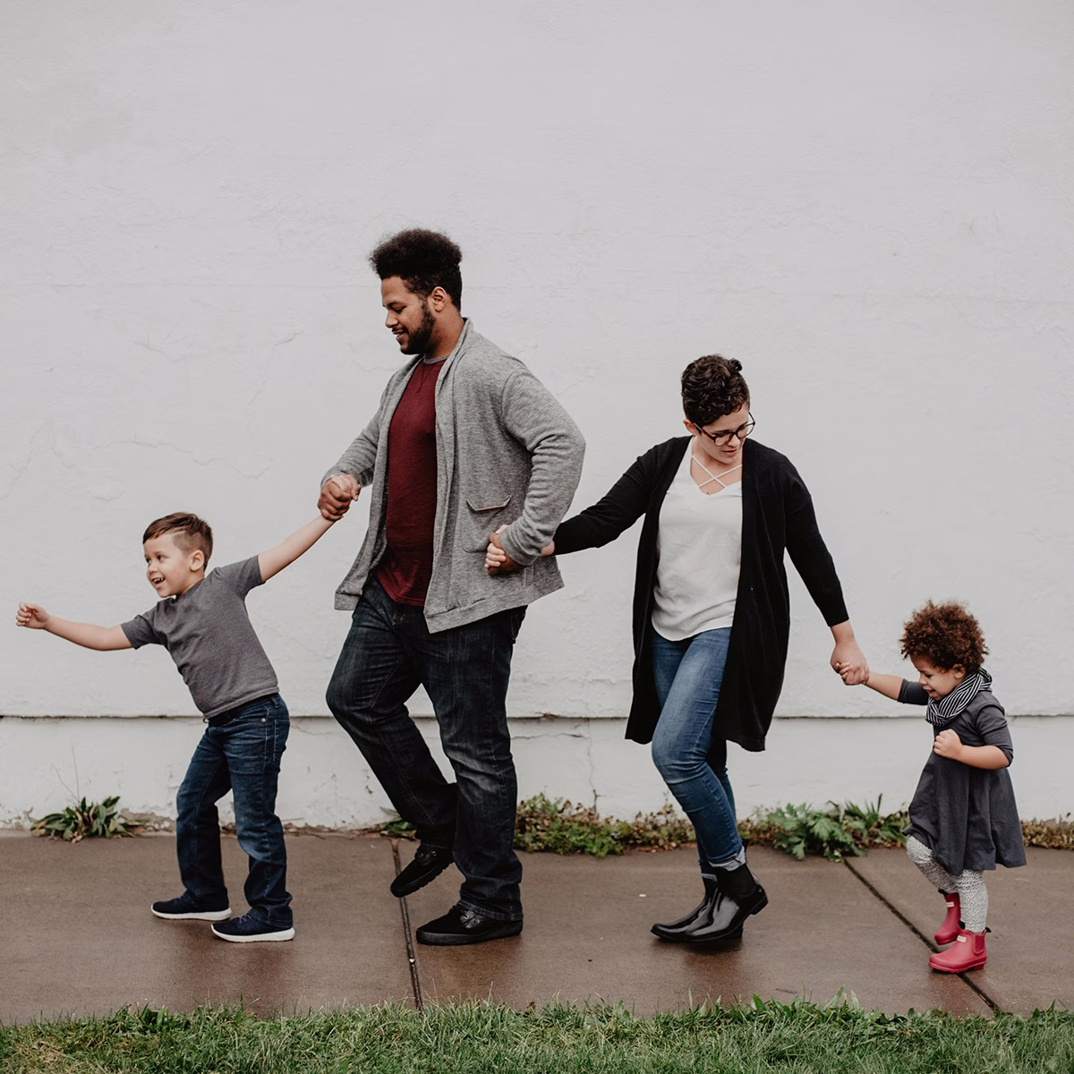 A family of four walking hand in hand along a sidewalk beside a white wall.