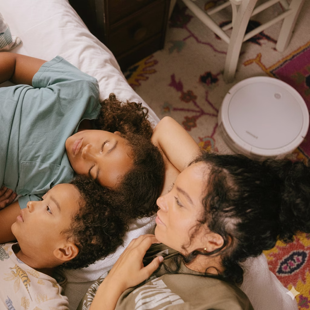 A parent and two children resting on a bed, with a white robot vacuum on the rug beside them.