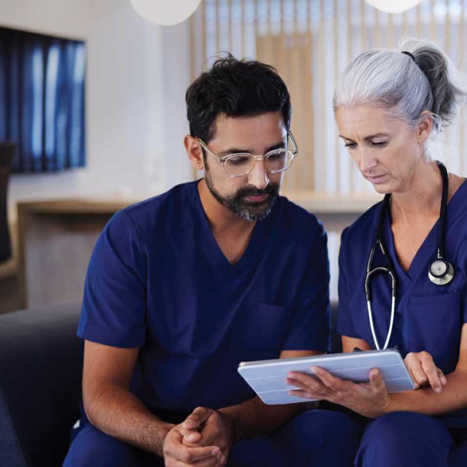 Two clinicians in navy scrubs sitting together, reviewing a patient record on a tablet.