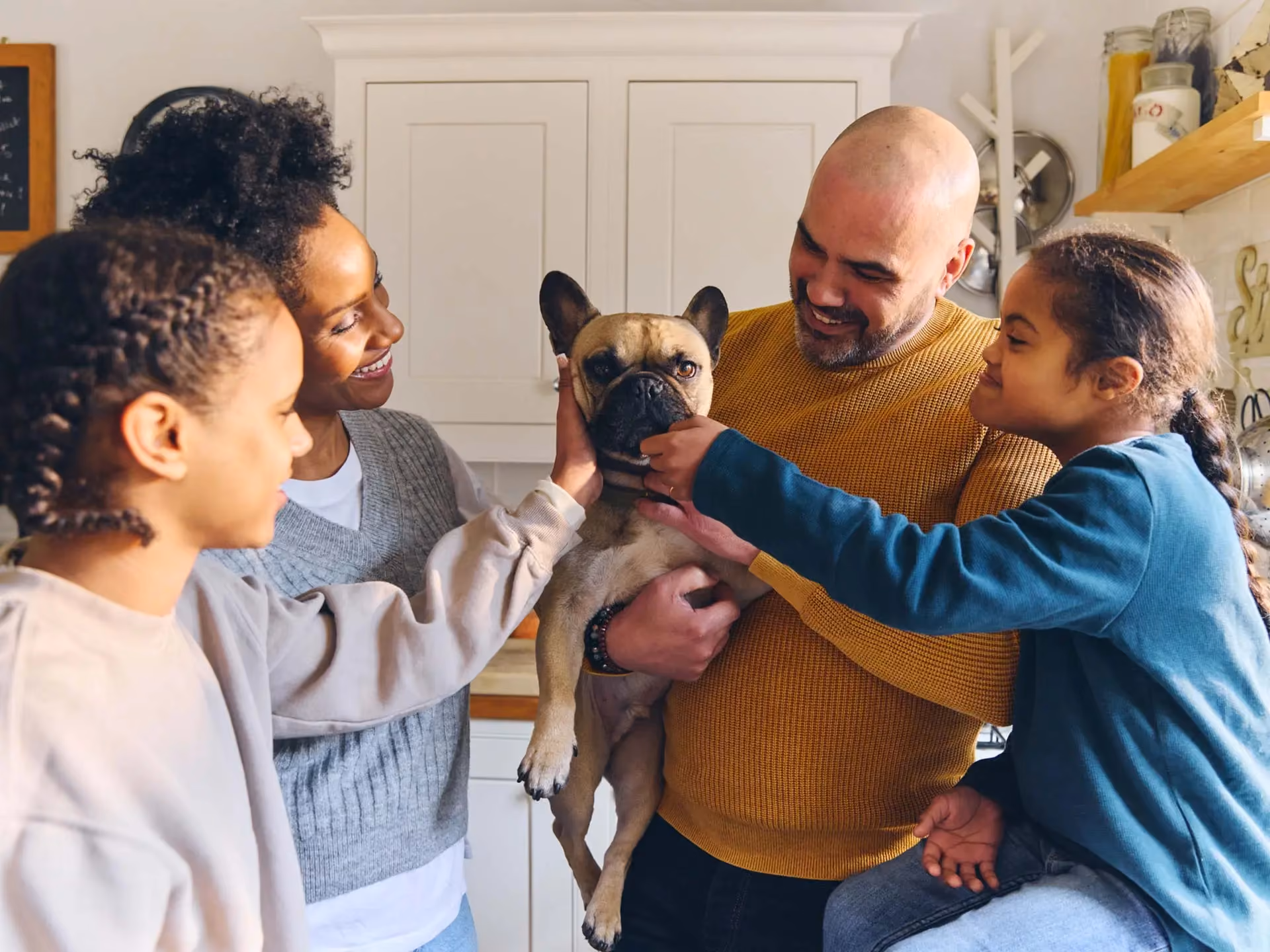 A family in a kitchen gathered around a French bulldog held in a man's arms.
