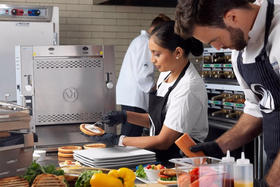 Kitchen staff preparing sandwiches on burger buns near a stainless steel commercial oven and prepped vegetables.