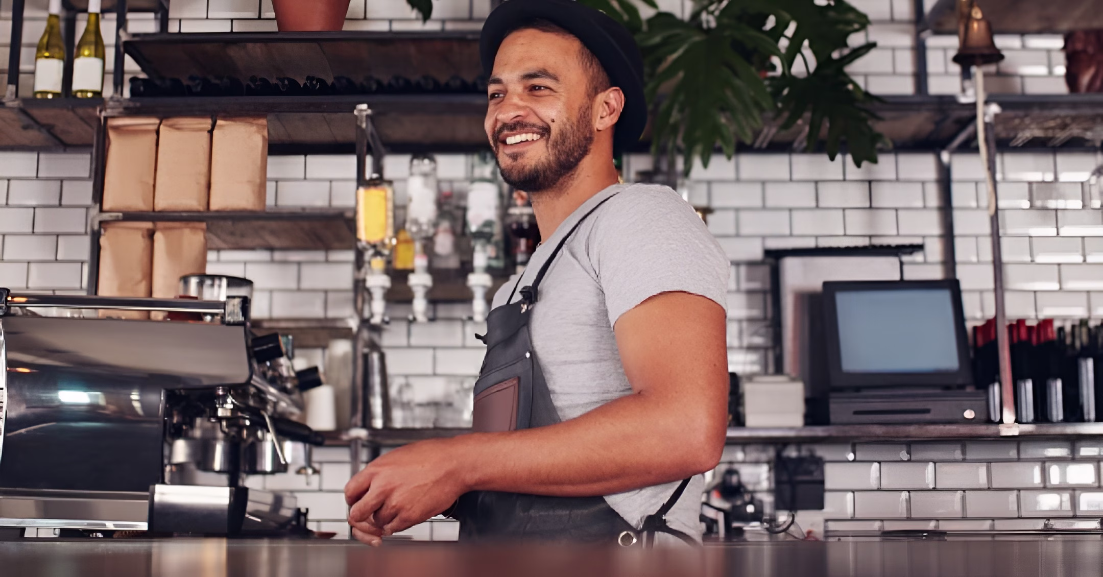 Smiling barista in a hat and apron standing behind a coffee shop counter with an espresso machine.