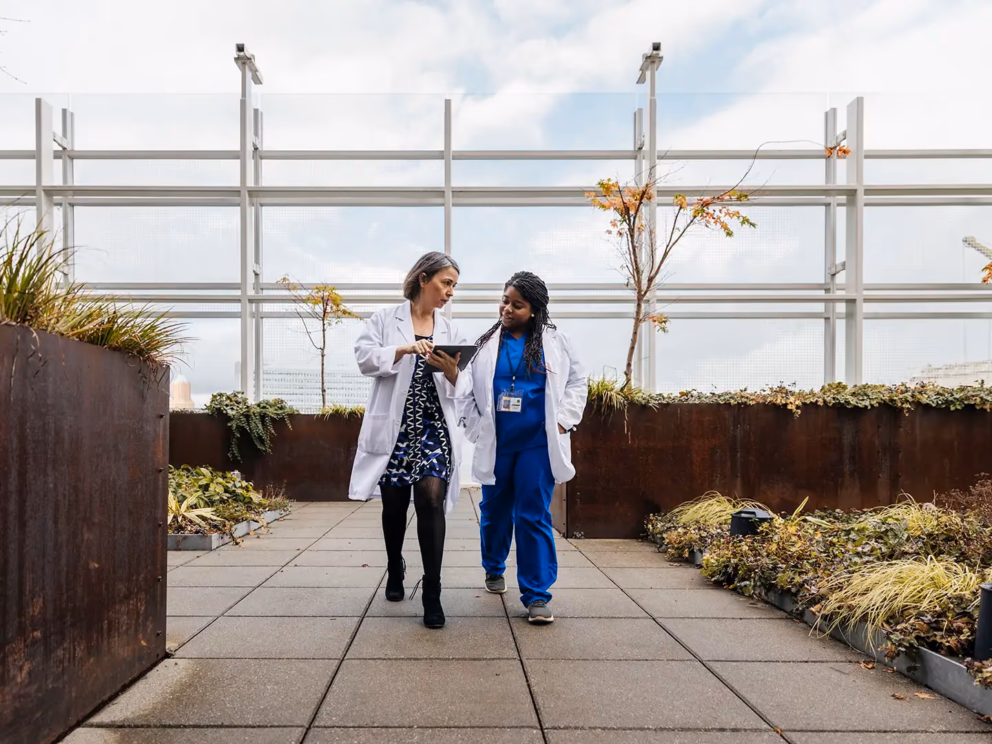Two women in white coats walk on a rooftop garden path, one holding a tablet, discussing its contents.