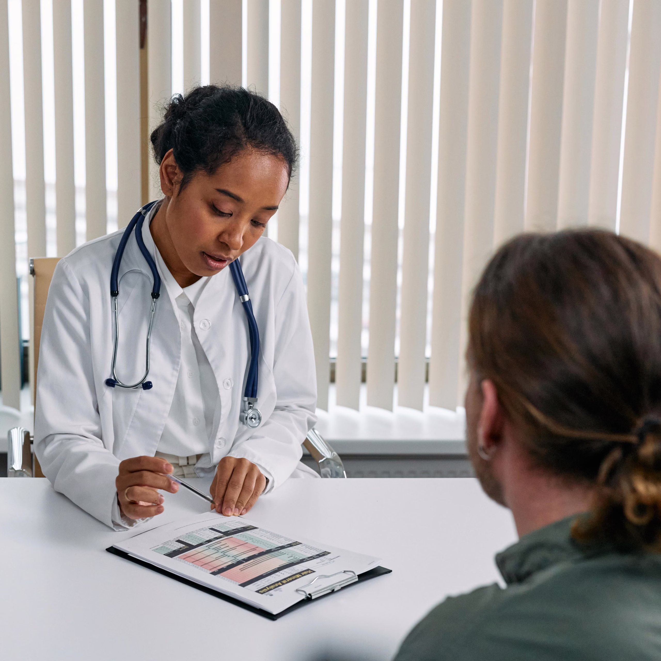 Doctor in white coat with stethoscope reviews a medical chart with a patient seated across the desk.