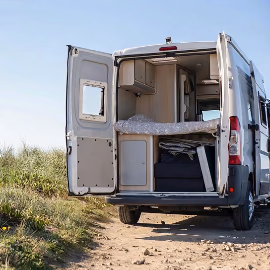 Camper van with rear doors open, showing interior bed and storage cabinets, parked on a dirt path.