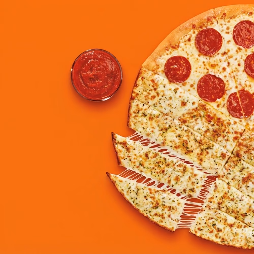 Half pepperoni, half cheese pizza cut into slices with cheesy garlic breadsticks, next to a cup of marinara sauce, on an orange background.