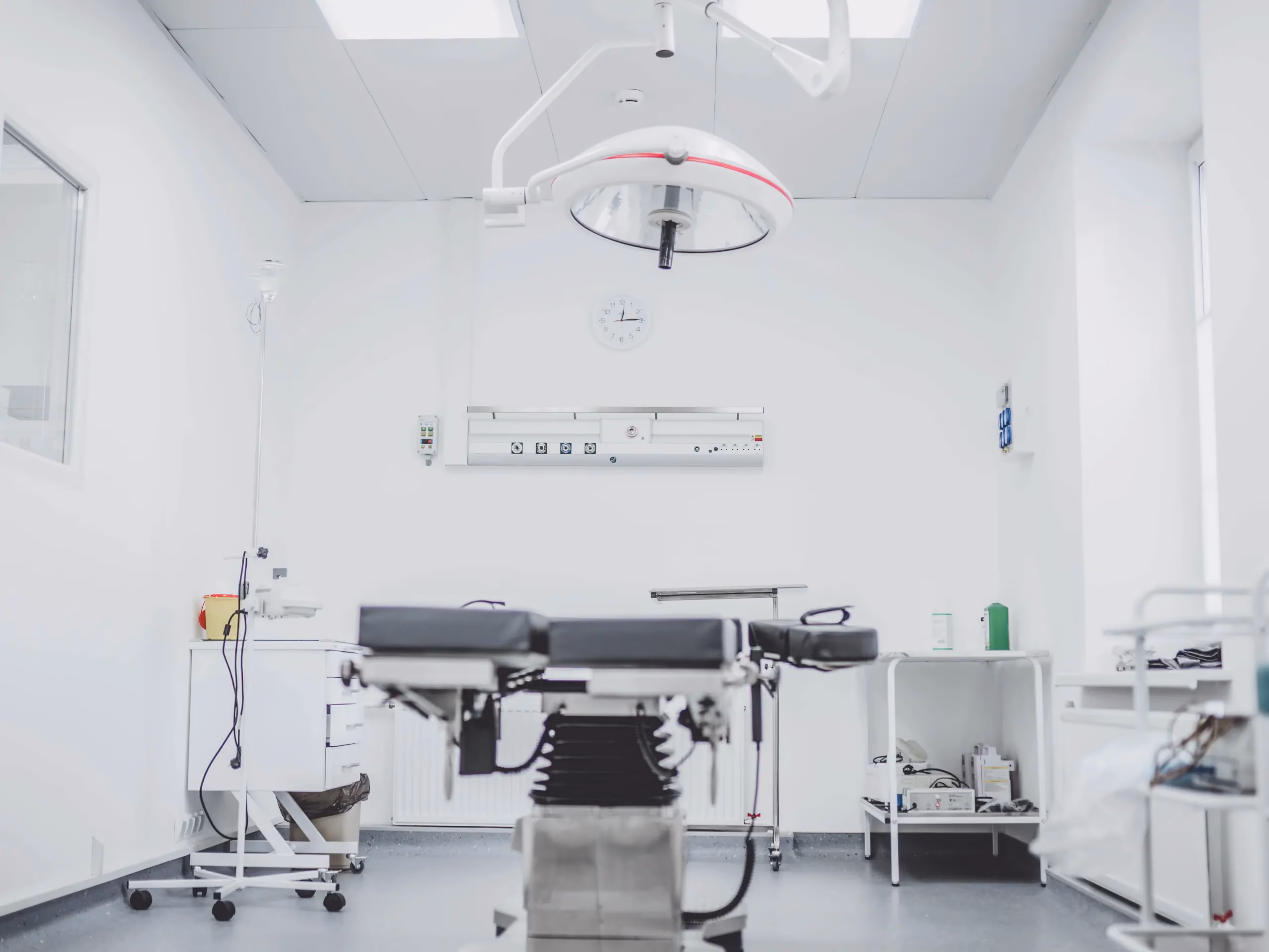Empty operating room with surgical table, overhead light, wall clock, IV pole, and equipment carts.