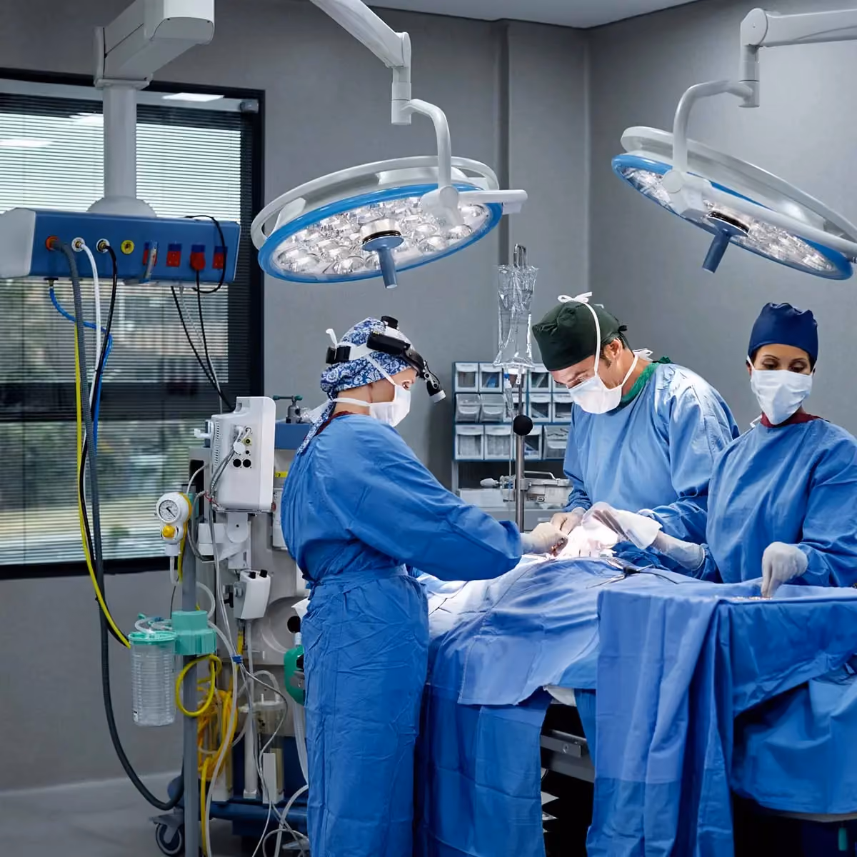 Three surgeons in blue scrubs and masks perform surgery under overhead lights in an operating room.