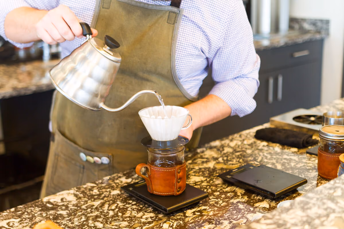 Person wearing an apron pours hot water from a gooseneck kettle over a coffee filter on a scale.