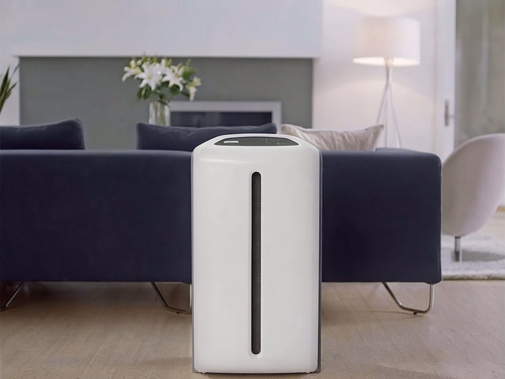 White air purifier standing on a wood floor in front of a dark blue sofa in a living room.