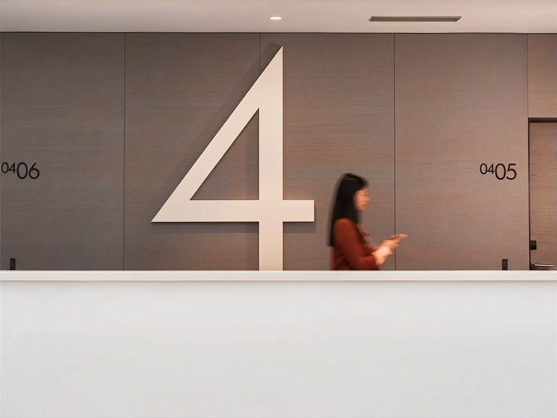 A large number 4 sign on a wall between room numbers 0406 and 0405, with a blurred woman walking past.