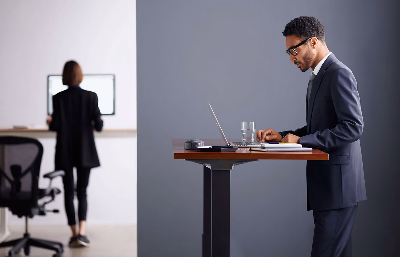 Man in a suit typing on a laptop at a standing desk, with a woman viewing a screen in the background.