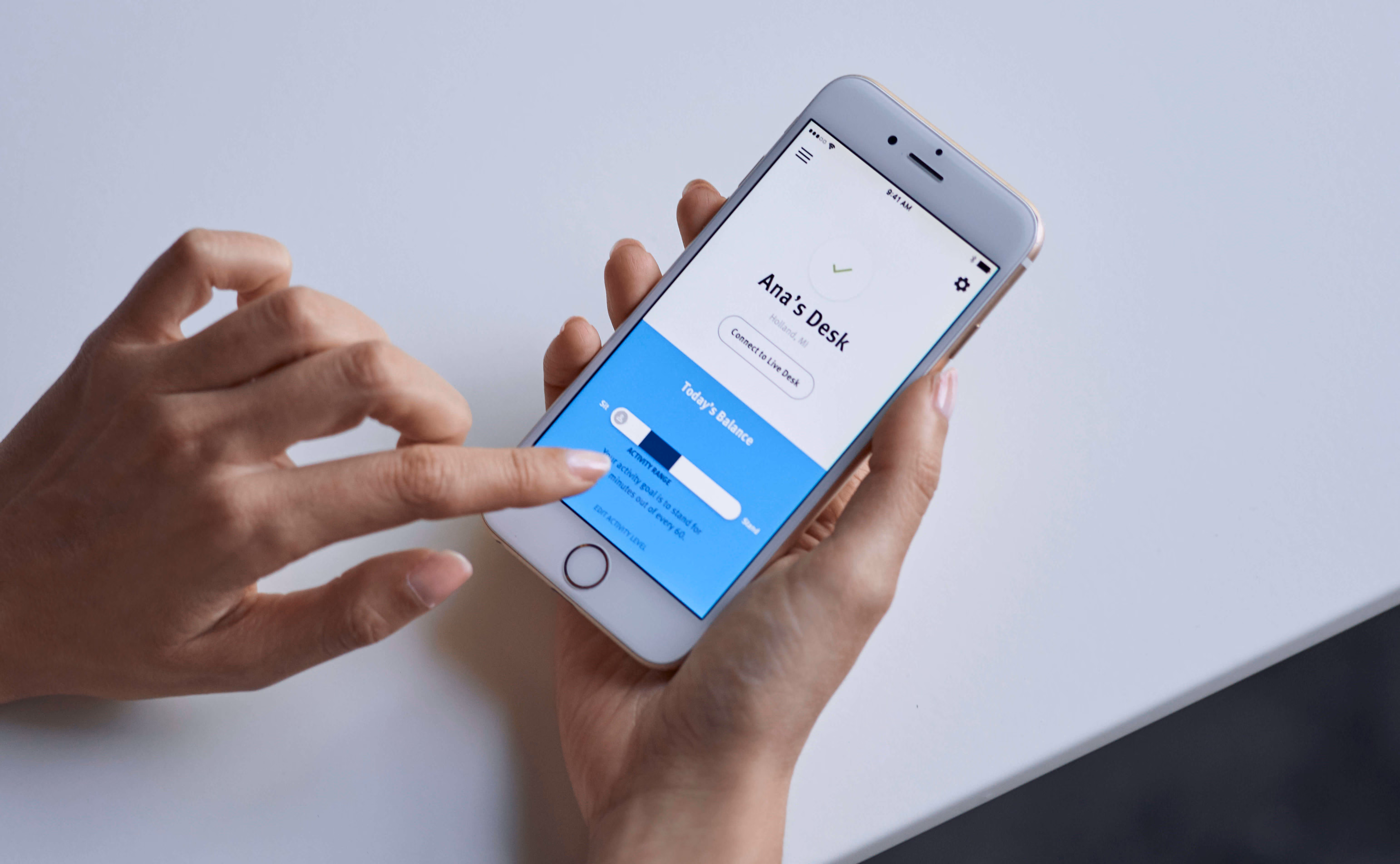 Hands holding a smartphone displaying a standing desk app labeled "Ana's Desk" with an activity range slider.