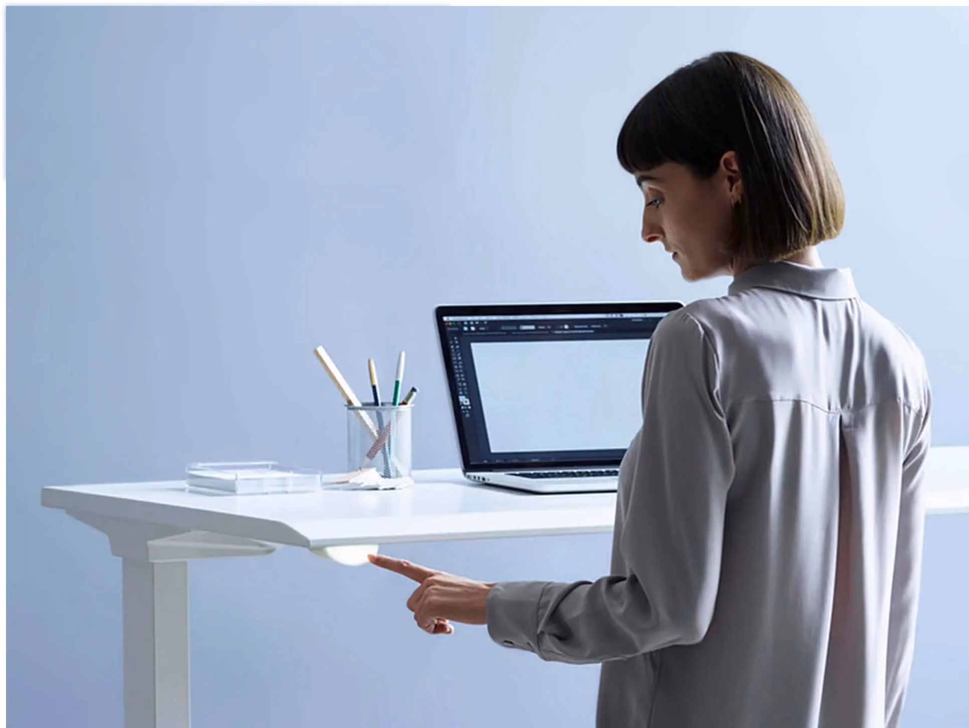 Woman in gray blouse touches a control button under a white standing desk, laptop and pencil cup on desktop.