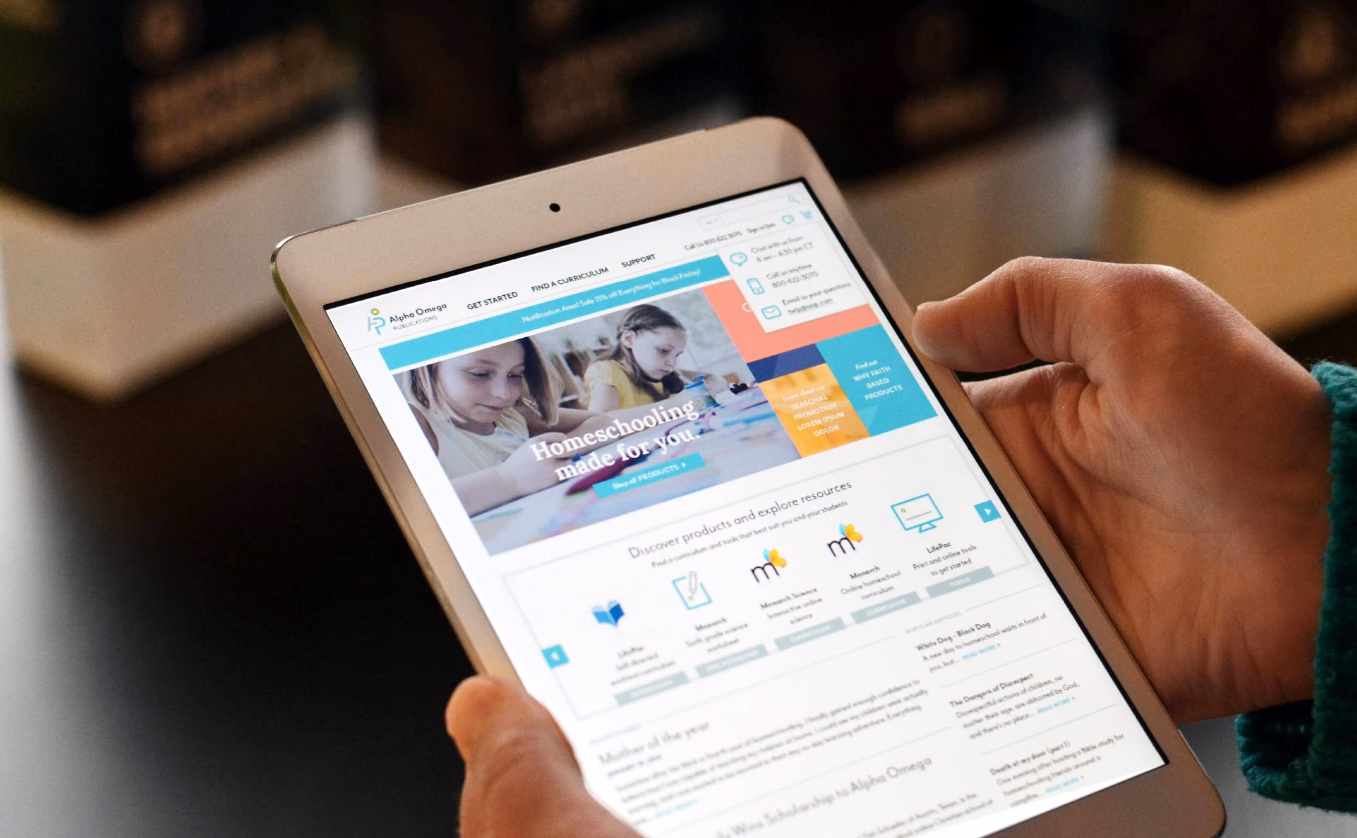 Hands holding a tablet displaying the Alpha Omega Publications homeschooling website.