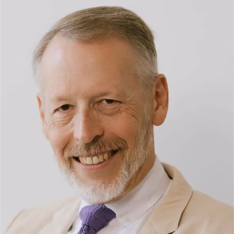 Headshot of a smiling older man with gray hair and a beard, wearing a beige blazer and purple knit tie.