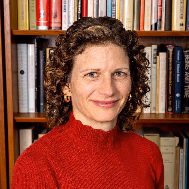 Woman with curly brown hair wearing a red turtleneck, smiling in front of a bookshelf.
