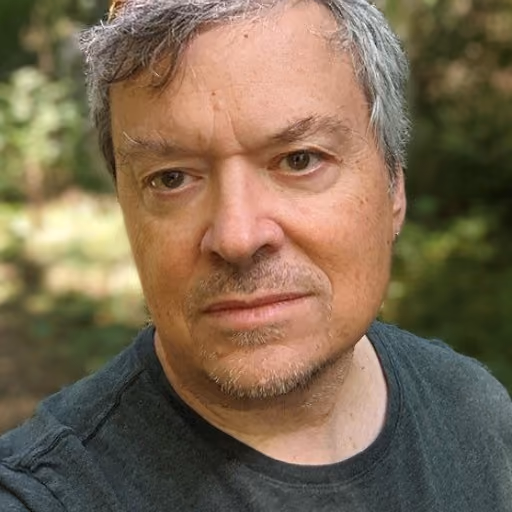 Portrait of a man with gray hair and a mustache, wearing a dark gray t-shirt, outdoors with trees in the background.