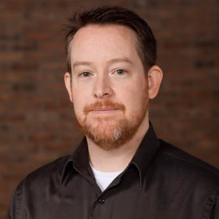Man with short brown hair and a beard wearing a dark button-up shirt, standing in front of a brick wall.