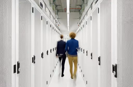 Two people walk down a narrow aisle between rows of server racks in a data center.