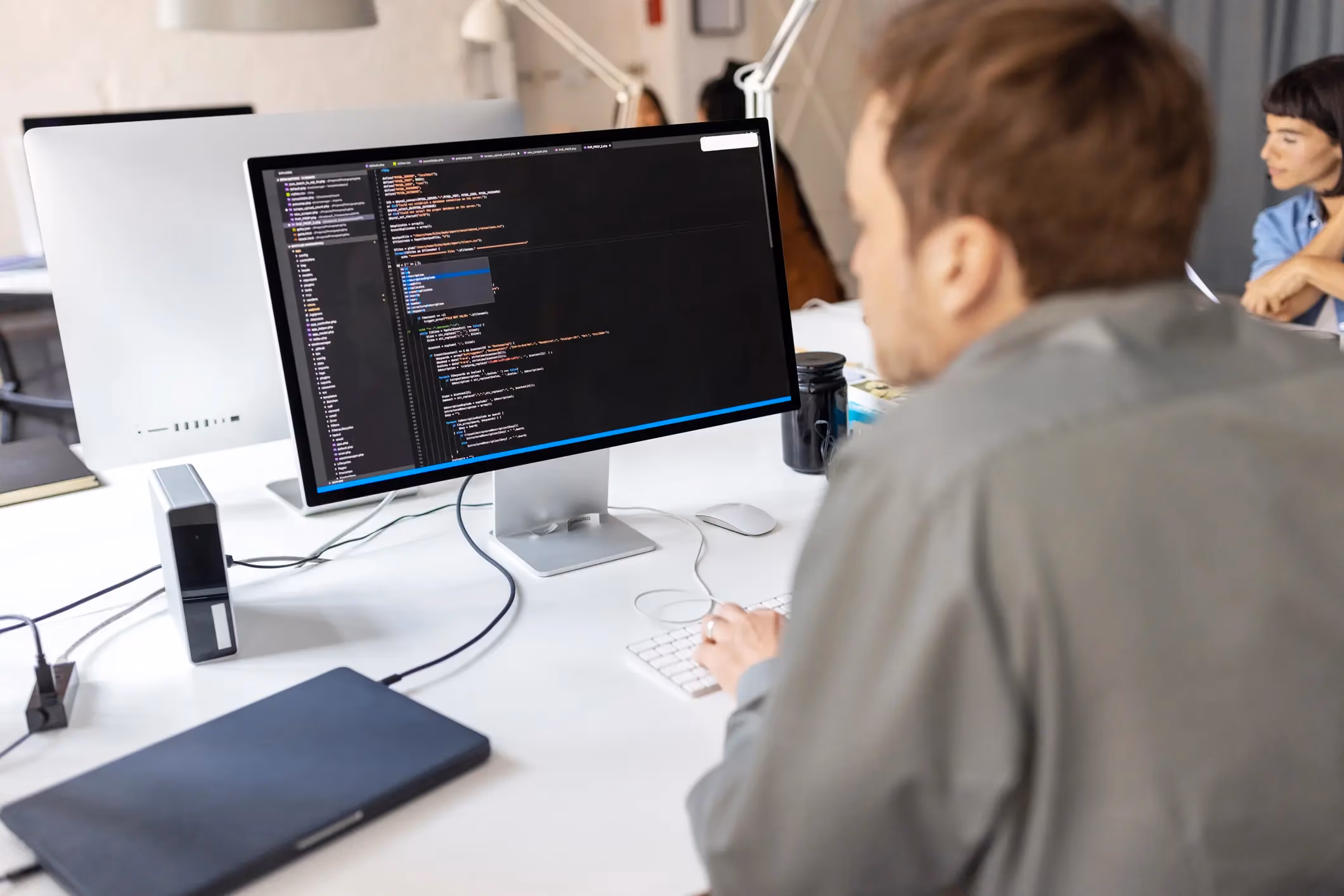 Man viewing code on a computer monitor at a white desk, with colleagues seated in the background.