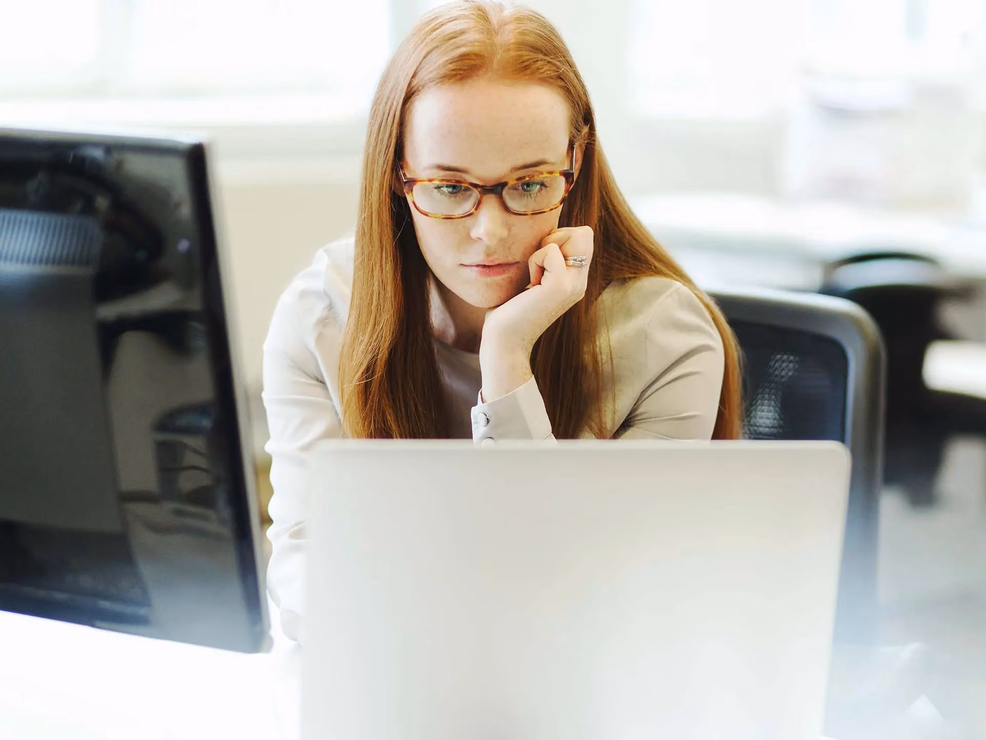 Woman with red hair and glasses sits at a desk, resting her chin on her hand while looking at a laptop screen.