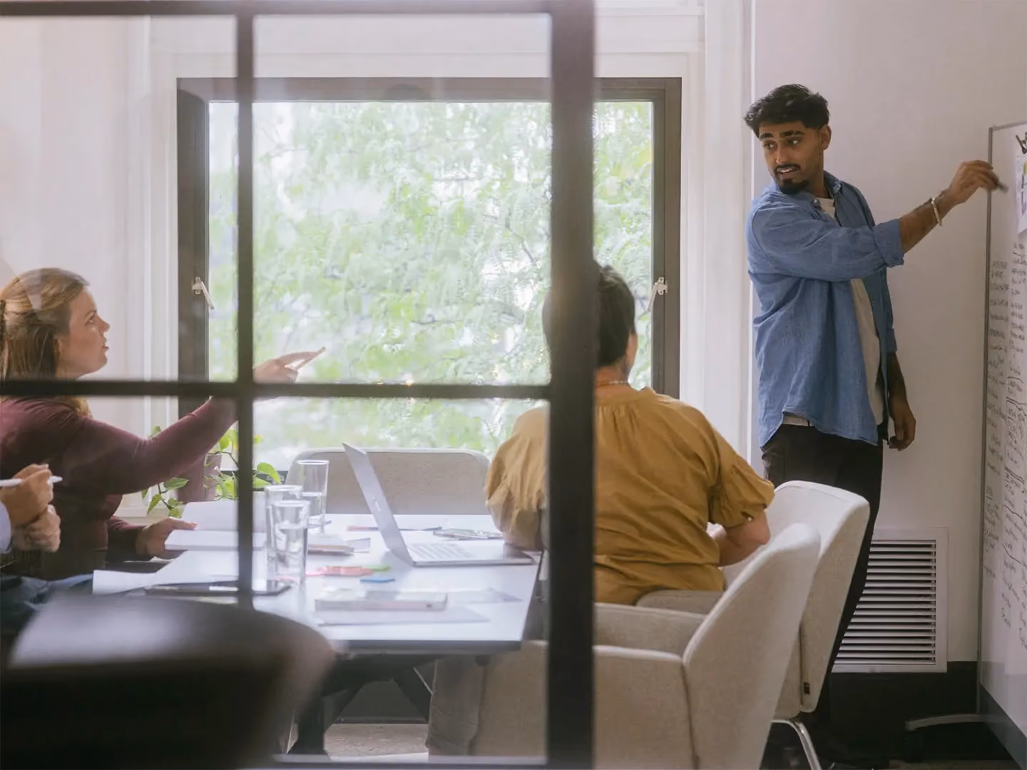 Man writing on a whiteboard while presenting to colleagues seated at a conference table near a window.